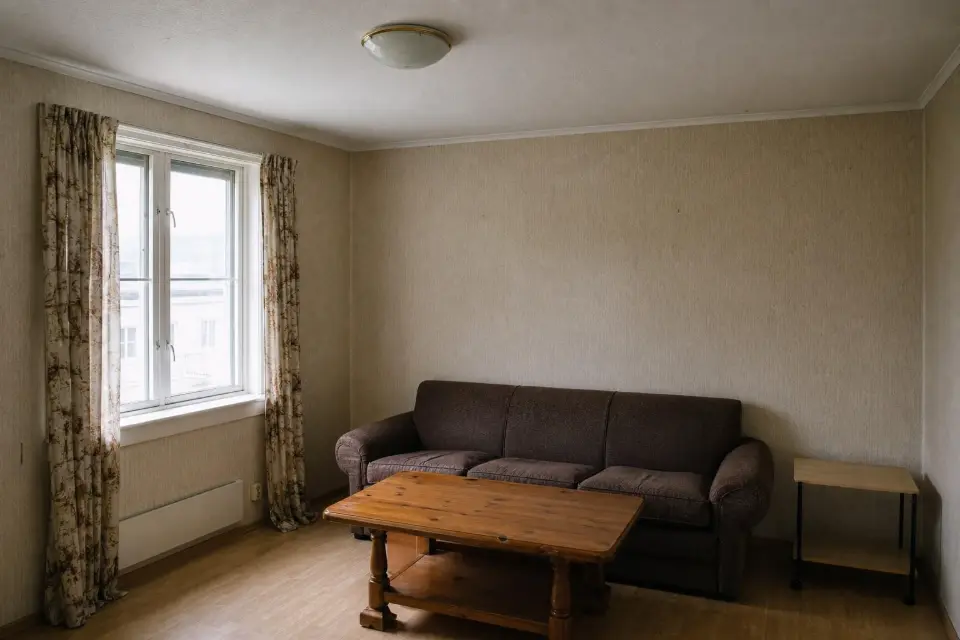 A tired 1990s living room with beige woodchip wallpaper, a worn brown sofa and a bulky pine coffee table.